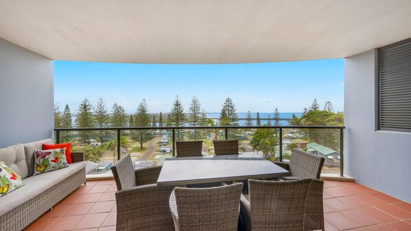A balcony with a table and chairs, a sofa, and a view of a coastal area with trees and buildings.