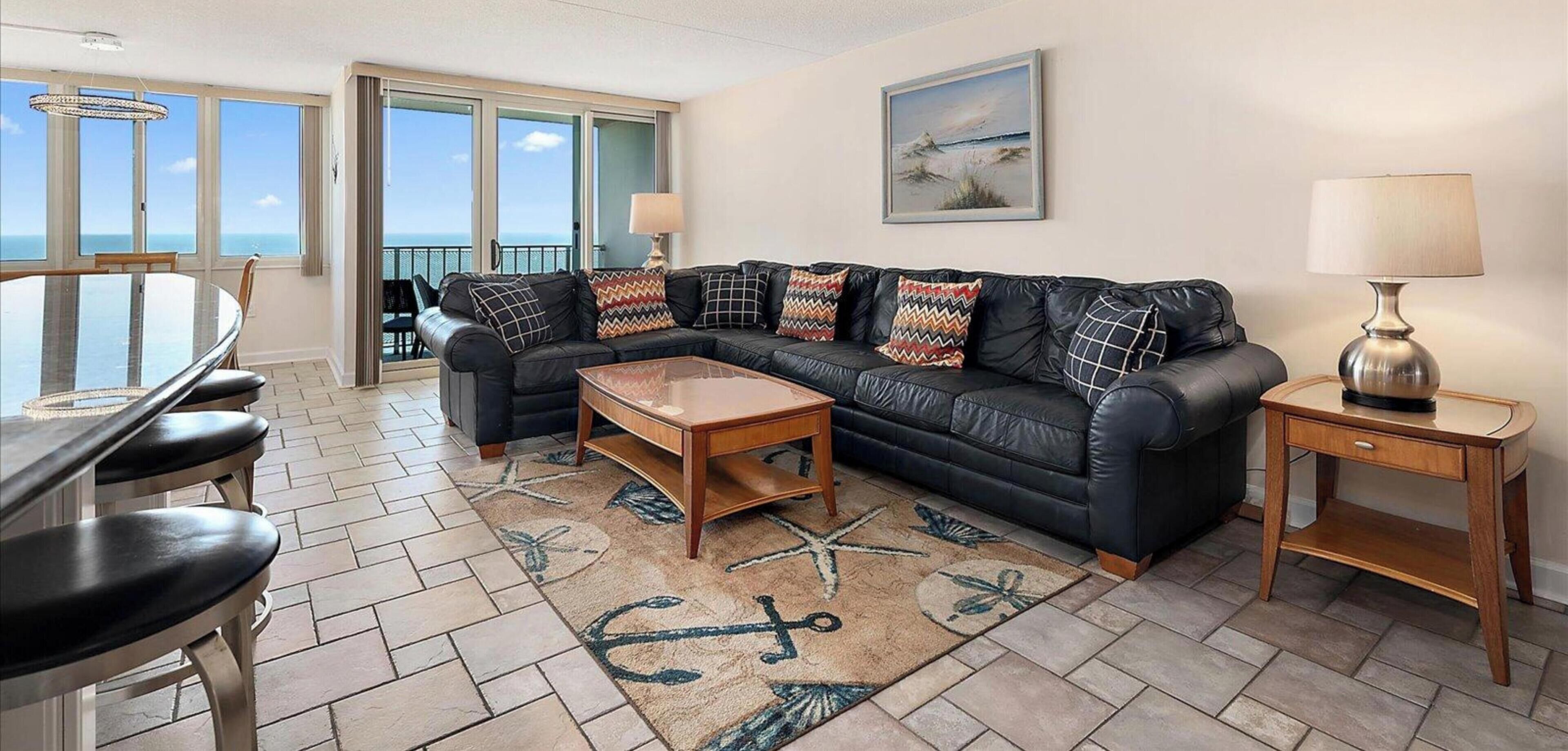 A living room with a black leather sofa, a coffee table, a dining table with chairs, and a view of the ocean through large windows.