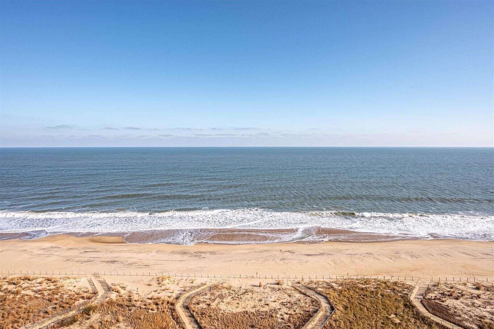 A beach with a clear view of the ocean, sandy terrain, and a fenced walking path.