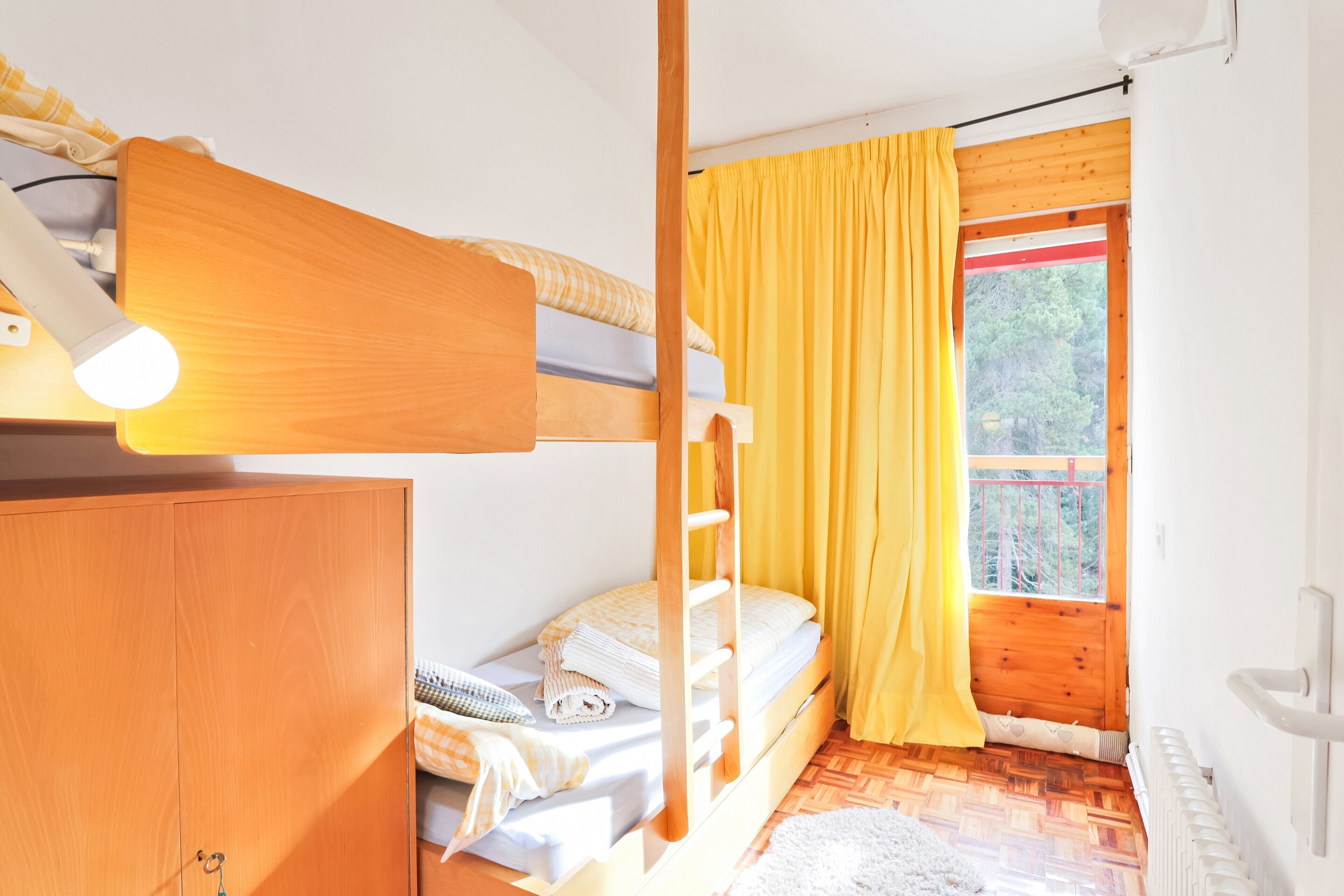 Une chambre avec un lit superposé, une armoire en bois, une fenêtre avec des rideaux jaunes et un radiateur.