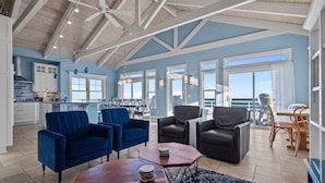 Amplia sala de estar con techo alto, paredes azules y ventanas grandes que ofrecen vista al océano. Hay dos sillones azules, una mesa de centro de madera y un sofá de cuero negro.