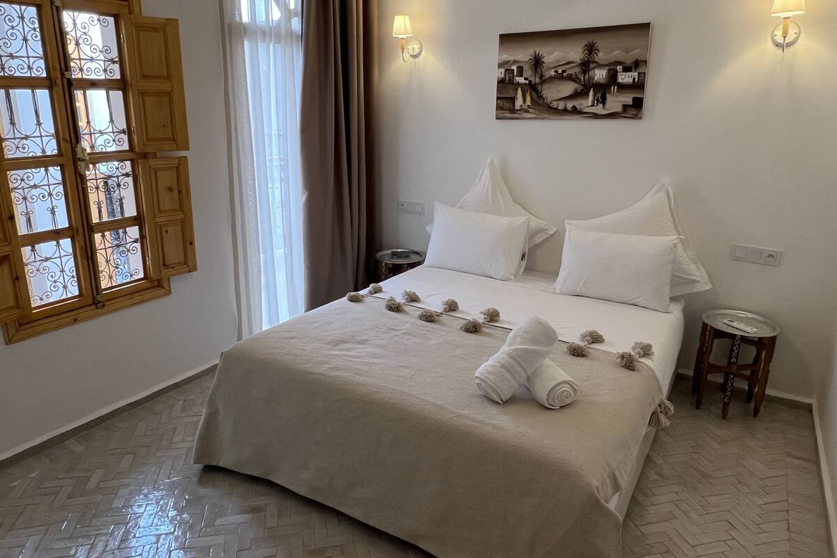 A neatly made bed with white linens and a rolled towel, a wall-mounted painting, two bedside lamps, and a window with decorative iron bars.