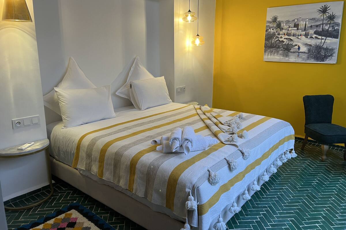 A hotel room with a bed, pillows, a striped blanket, a towel, a chair, and a painting on the wall.