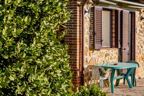 A brick building with a window and shutter, a table with chairs, and a green bush in the foreground. - Farmhouse "Le Gerlette - La Capanna" with Shared Pool & Wi-Fi (Braccagni)
