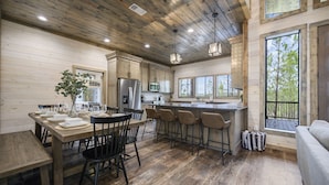 A spacious kitchen with wooden floors, a dining area with a large table and chairs, and a bar area with stools. - NEW Cozy Lux Cabin! *VIEWS* Hot Tub,Wi-Fi-Sleeps 8 (Broken Bow)