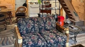 A cozy cabin interior with a wooden floor, a floral sofa, a dining table with chairs, a kitchen area with a stove, and a wooden staircase.
