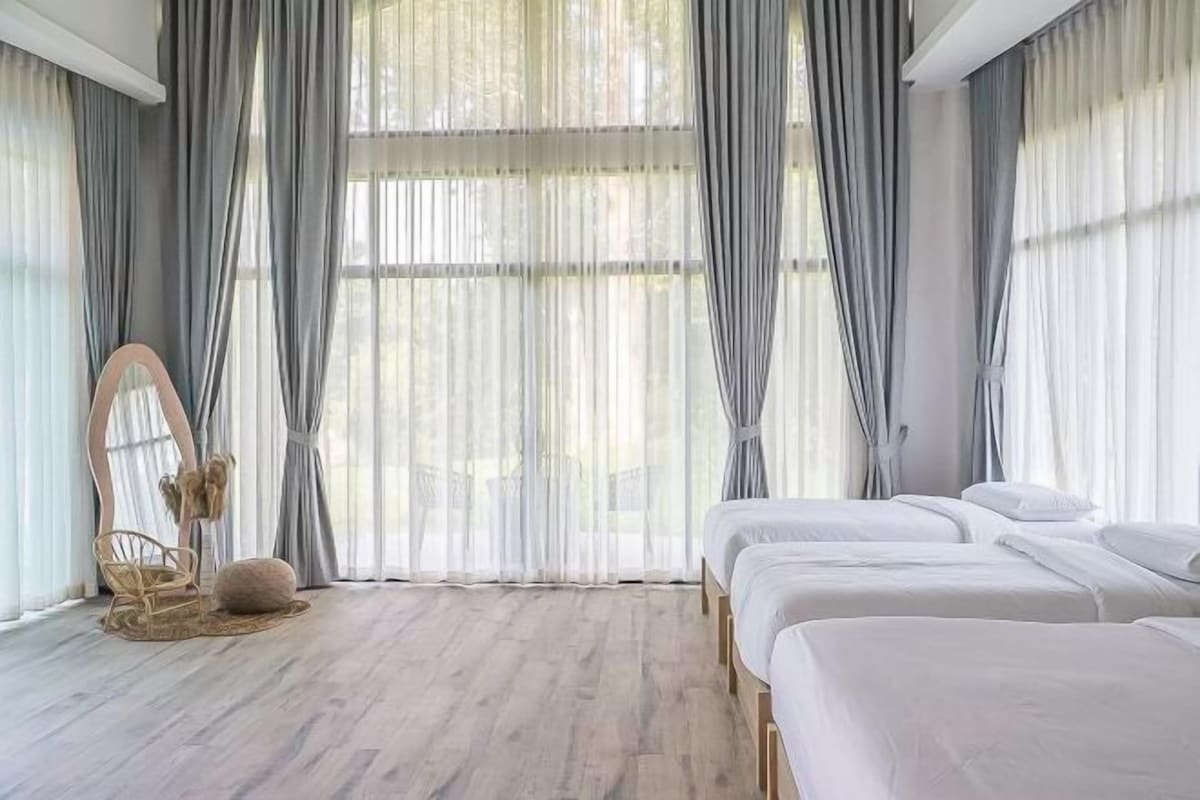A room with three beds, a large window with sheer curtains, a wooden floor, and a round wooden bench.