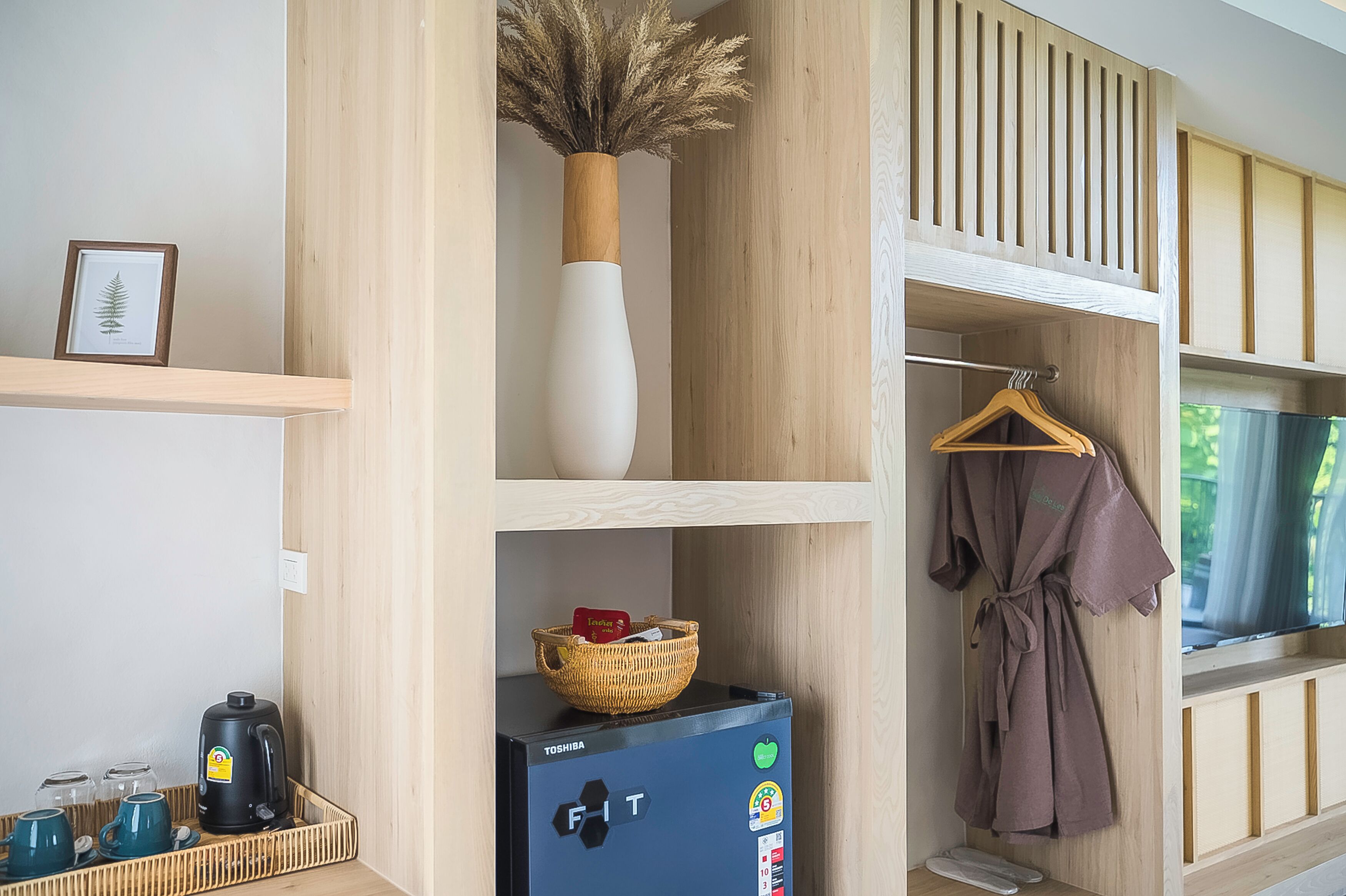 Une pièce bien rangée, avec un réfrigérateur Toshiba noir, un vase blanc contenant des plantes séchées, un tableau encadré et un porte-vêtements en bois sur lequel est posé un peignoir.