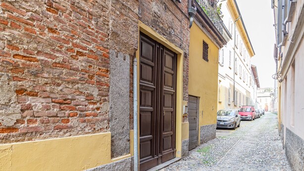 A narrow cobblestone street with a brick building on the left and a yellow building on the right. There are parked cars along the street. - VickyLù (Pavia)