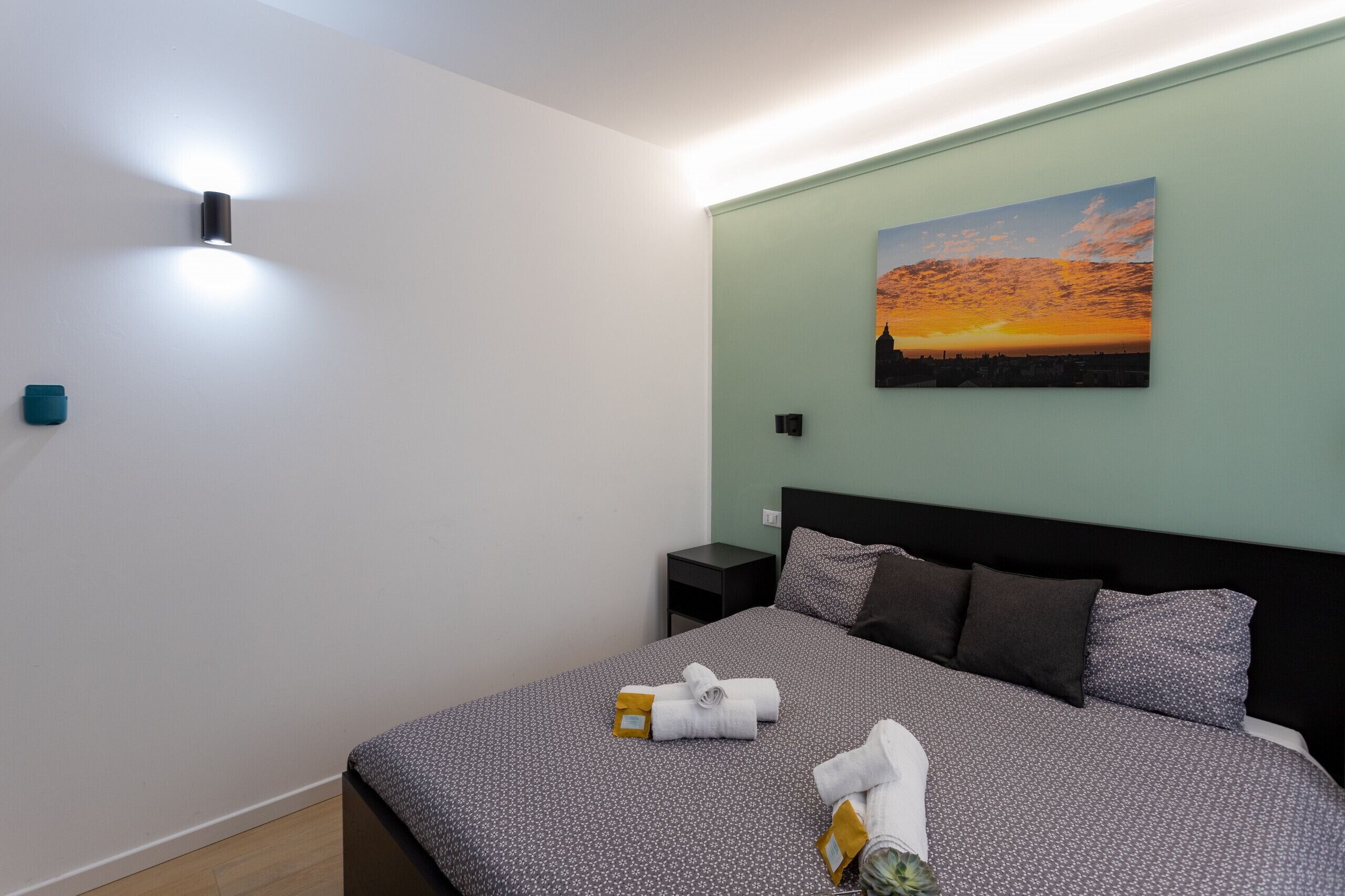 A neatly made bed with a patterned bedspread, a painting of a sunset on the wall, and a wall-mounted light fixture.