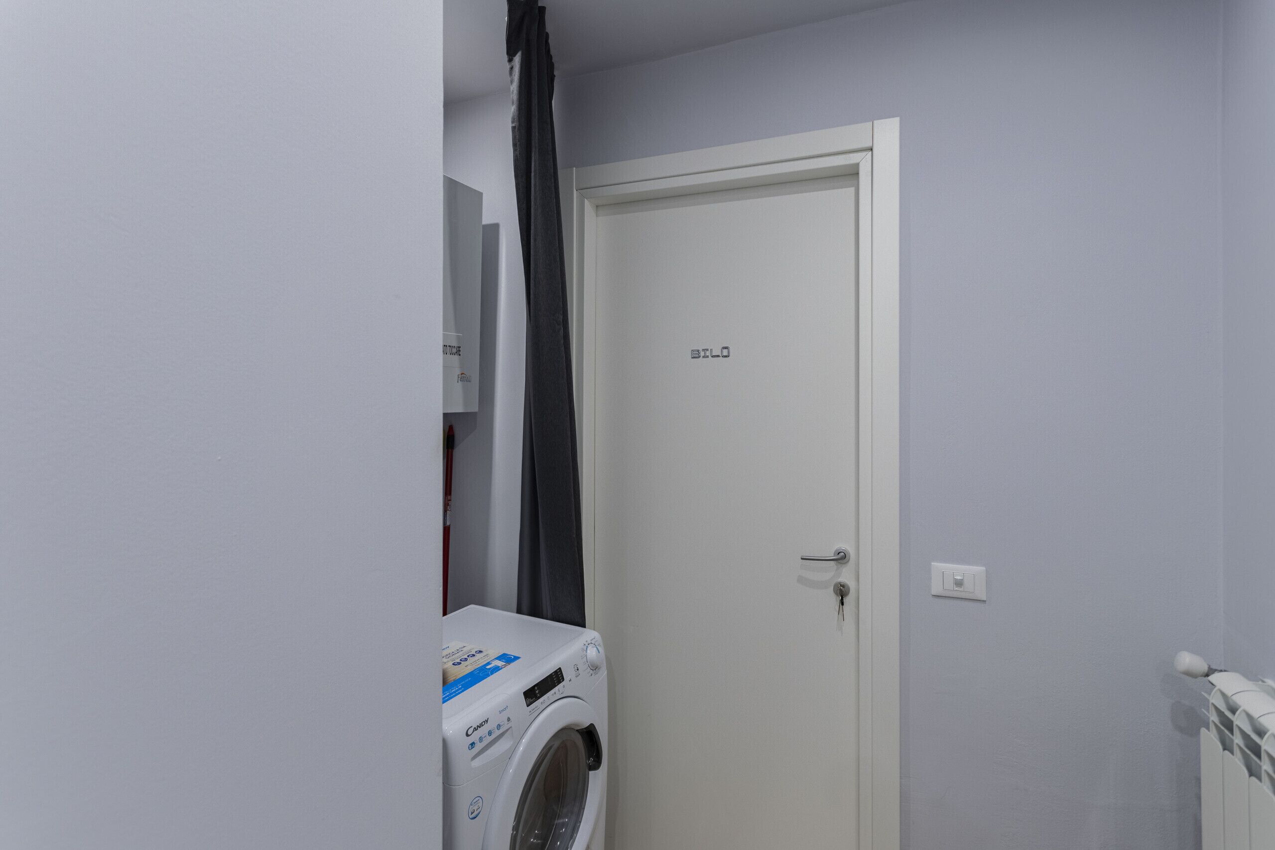 Une petite buanderie équipée d’un lave-linge, d’une porte portant l’inscription « BILO » et d’un radiateur.