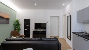 Un salon moderne avec un canapé en cuir noir, un téléviseur à écran plat, une bibliothèque et un coin repas équipé d’un évier et d’un miroir.