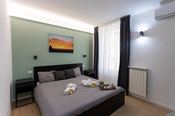 Un lit bien fait, recouvert d’une couvre-lit à motifs, une petite table de chevet, un climatiseur fixé au mur et un tableau représentant un coucher de soleil.