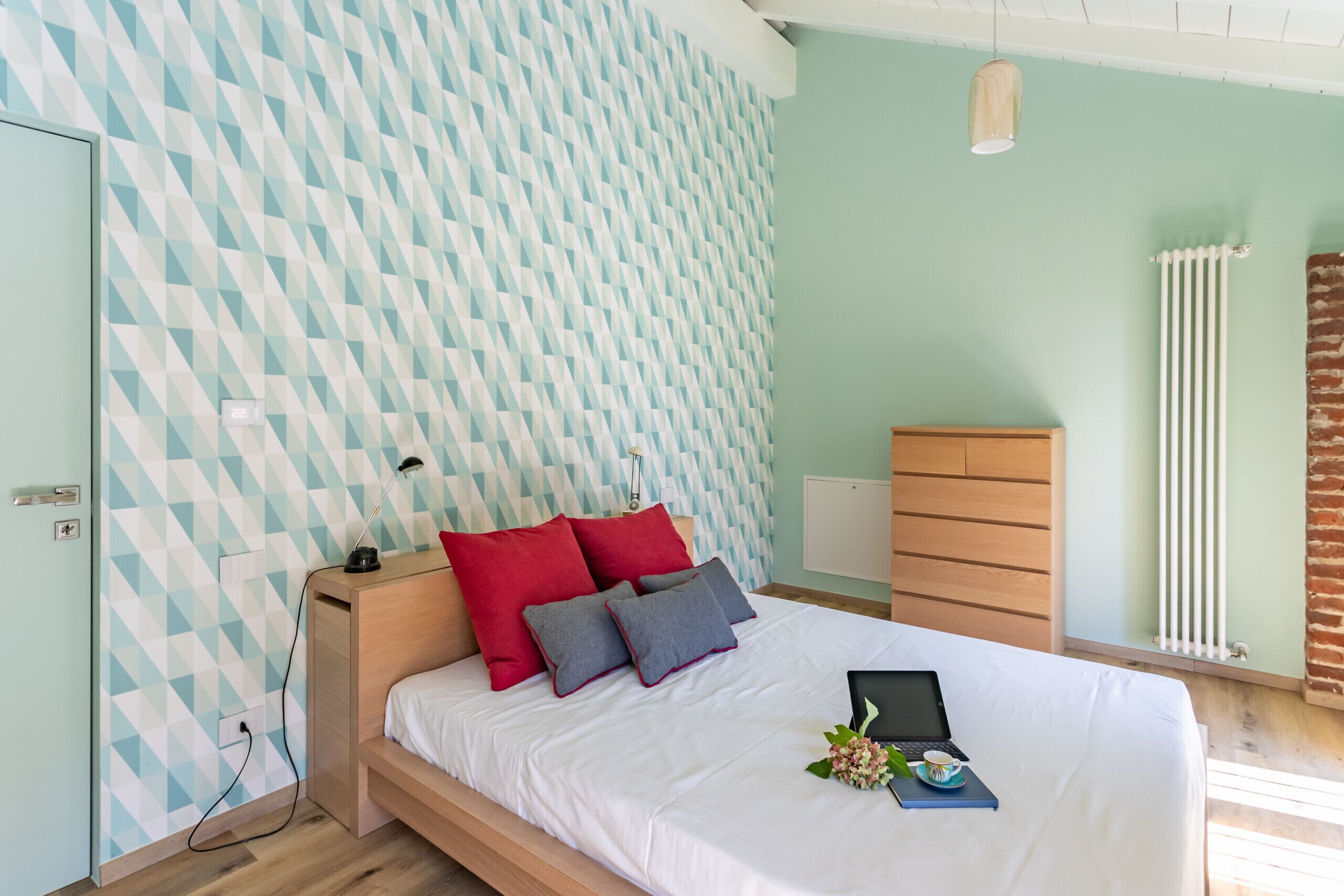Een netjes opgemaakt bed met een wit laken, rode en grijze kussens, en een laptop op het bed.