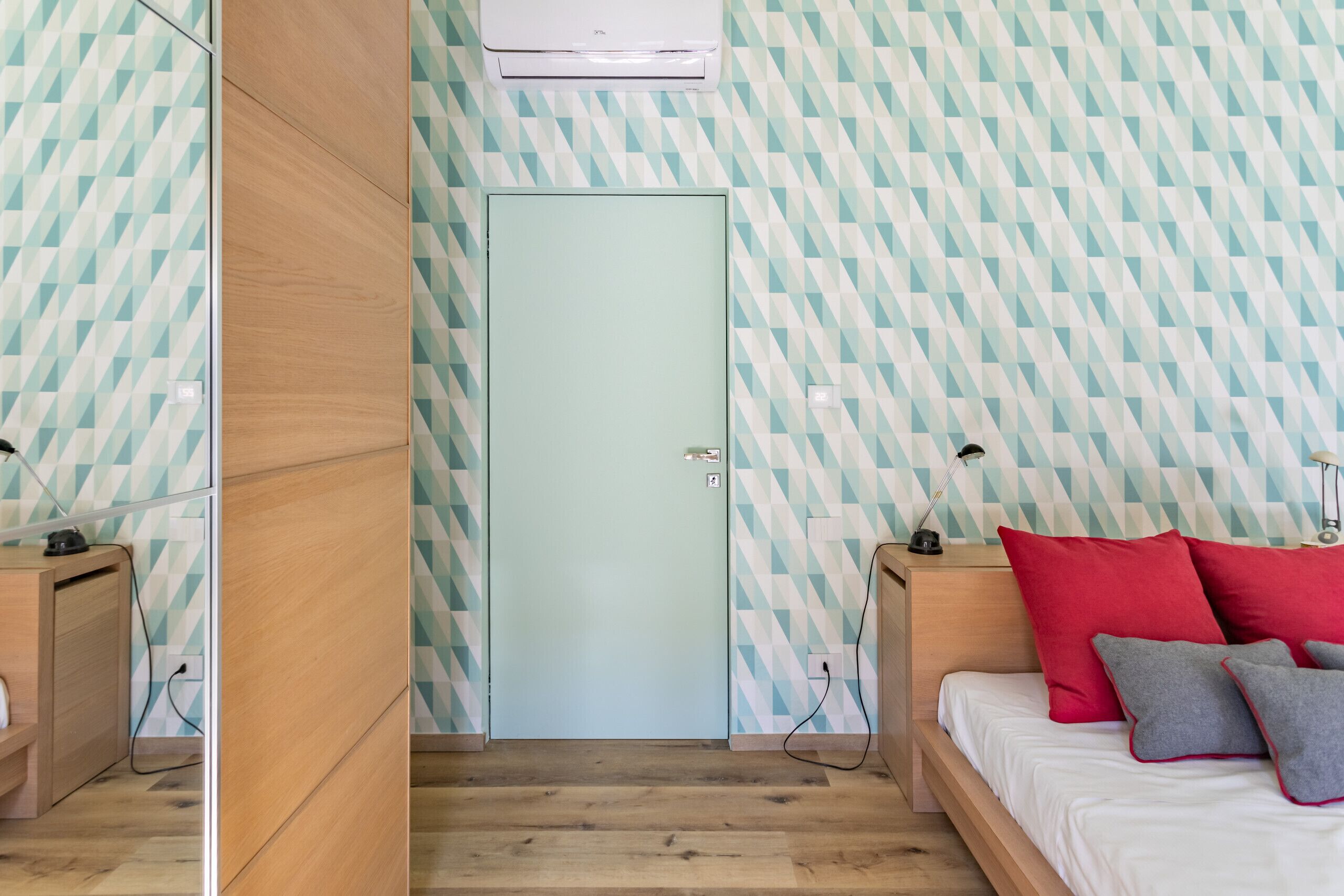 Une pièce avec un mur orné d’un motif géométrique, une porte bleu clair, un lit avec des coussins rouges et gris, et des meubles en bois.