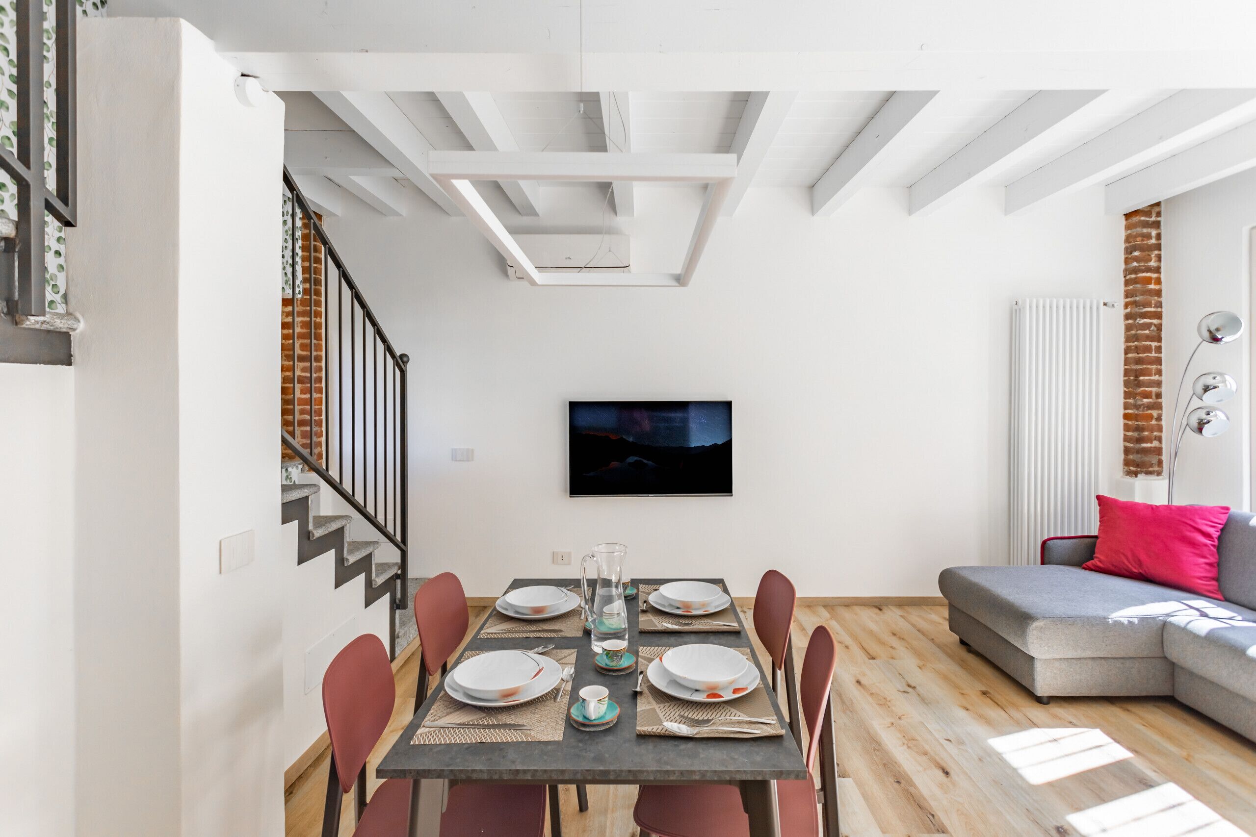 A dining area with a table set for six, a TV mounted on the wall, and a staircase leading to another level.