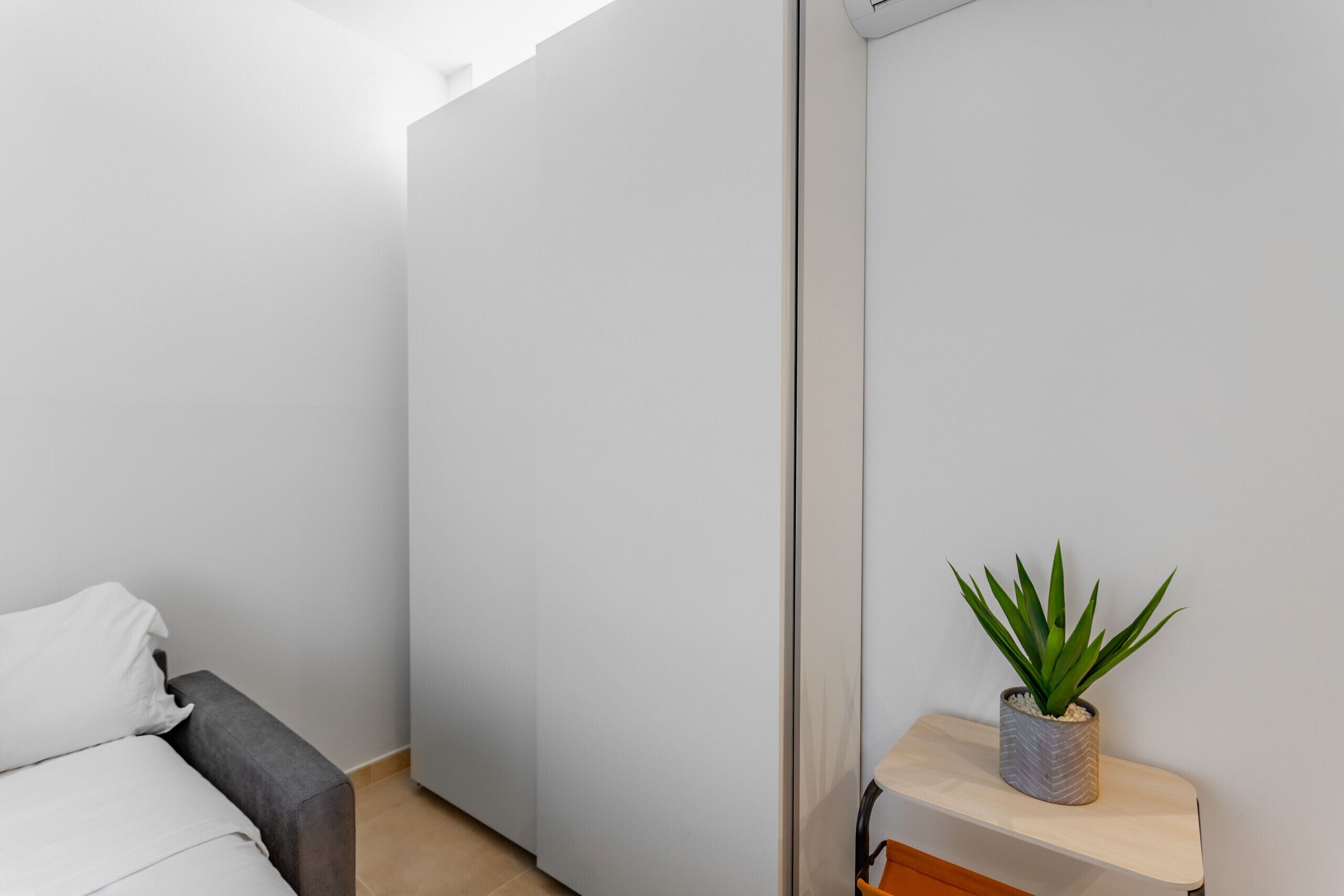 Ett minimalistiskt rum med en säng, en grå soffa, en liten hylla med en växt och en vit dörr.