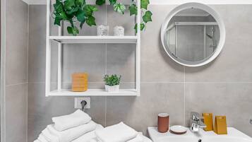 A bathroom with a white sink, a round mirror, a shelf with plants, and neatly folded towels.