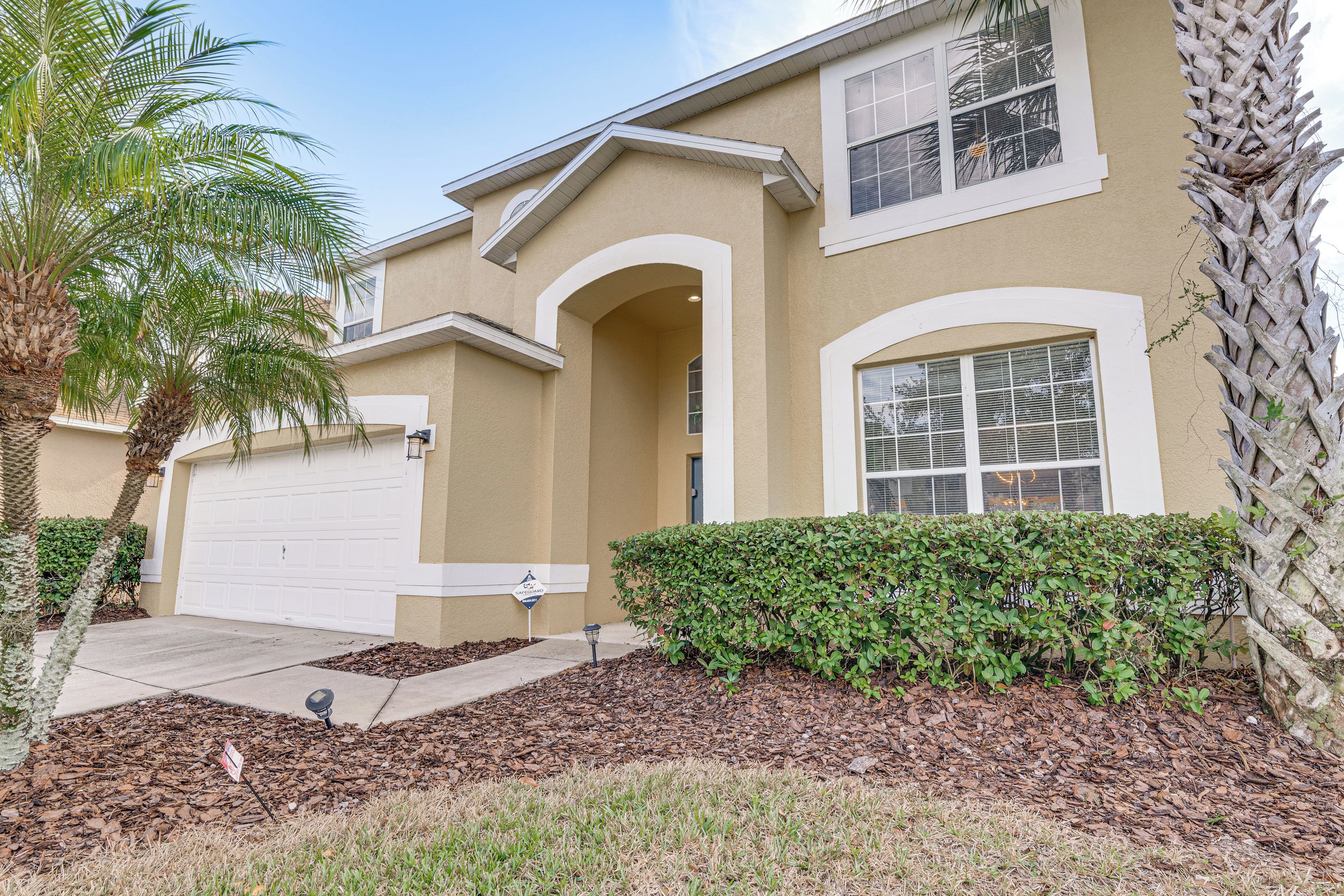 A two-story house with a beige exterior, white trim, and a garage. There are palm trees and a well-maintained lawn with mulch.