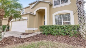 A two-story house with a beige exterior, white trim, and a garage. There are palm trees and a well-maintained lawn with mulch. - 6BR Disney Villa Pool, Spa & Game Room (Kissimmee)