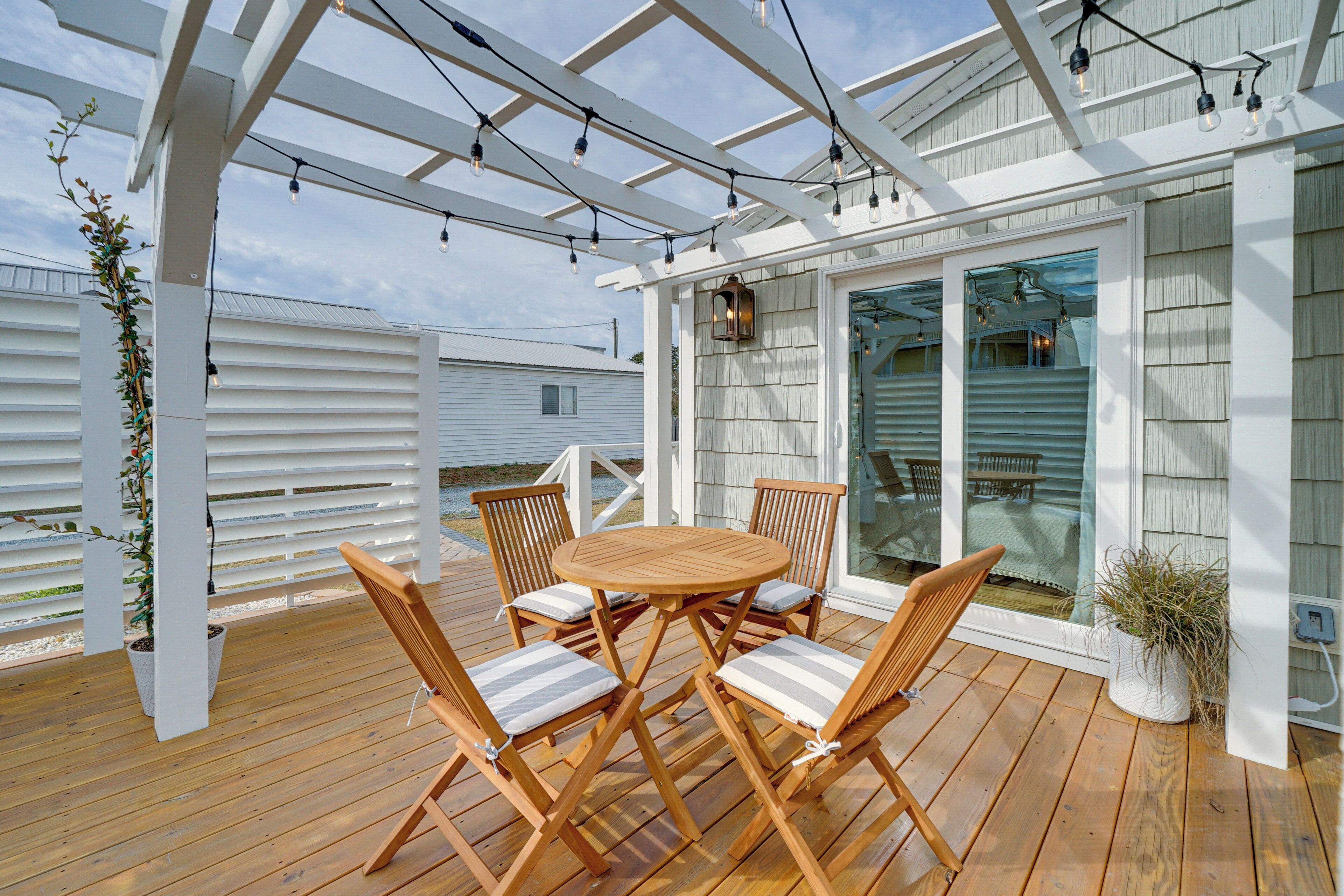 A wooden deck with a round table and chairs, a pergola with string lights, and a view of a house through glass doors.
