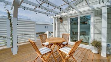 A wooden deck with a round table and chairs, a pergola with string lights, and a view of a house through glass doors.