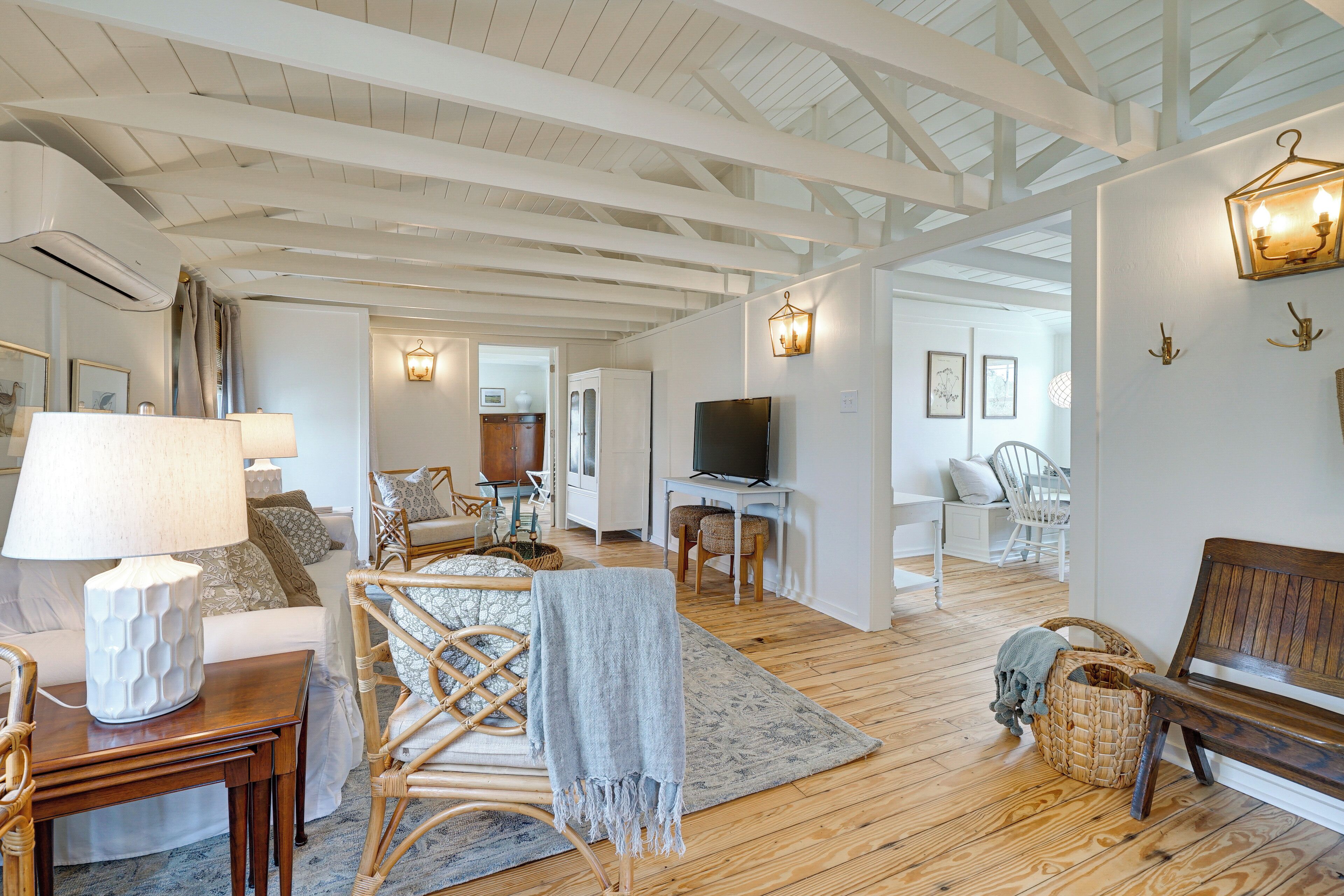 A spacious living room with wooden floors, a white couch, a wooden bench, wicker chairs, a TV, and a fireplace.