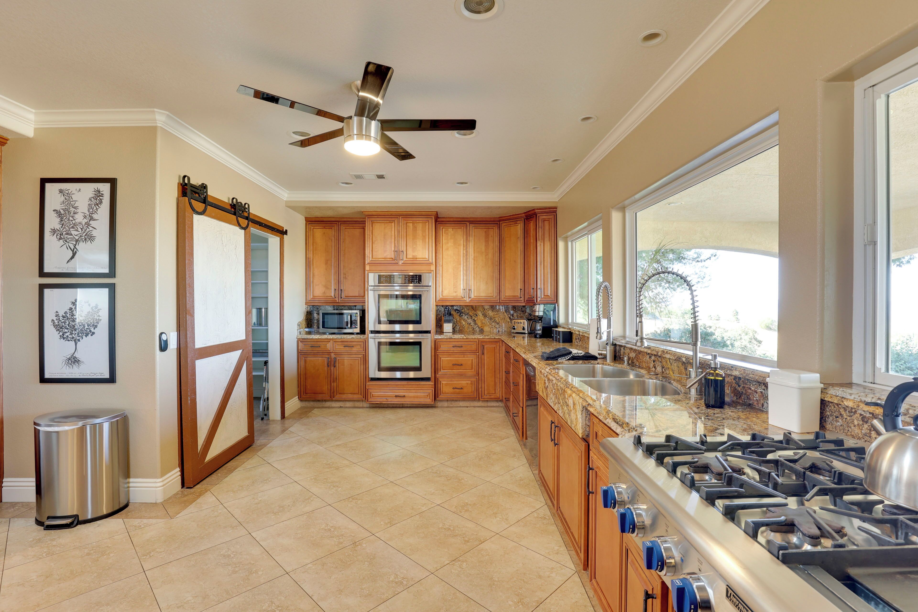 A modern kitchen with wooden cabinets, stainless steel appliances, a large window, and a ceiling fan.