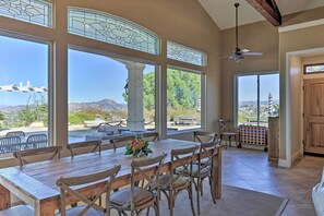 A dining area with a large wooden table and chairs, large windows with decorative glass, and a view of mountains and greenery. - Charming Temecula Home w/ Hot Tub, Mountain Views! (Temecula)