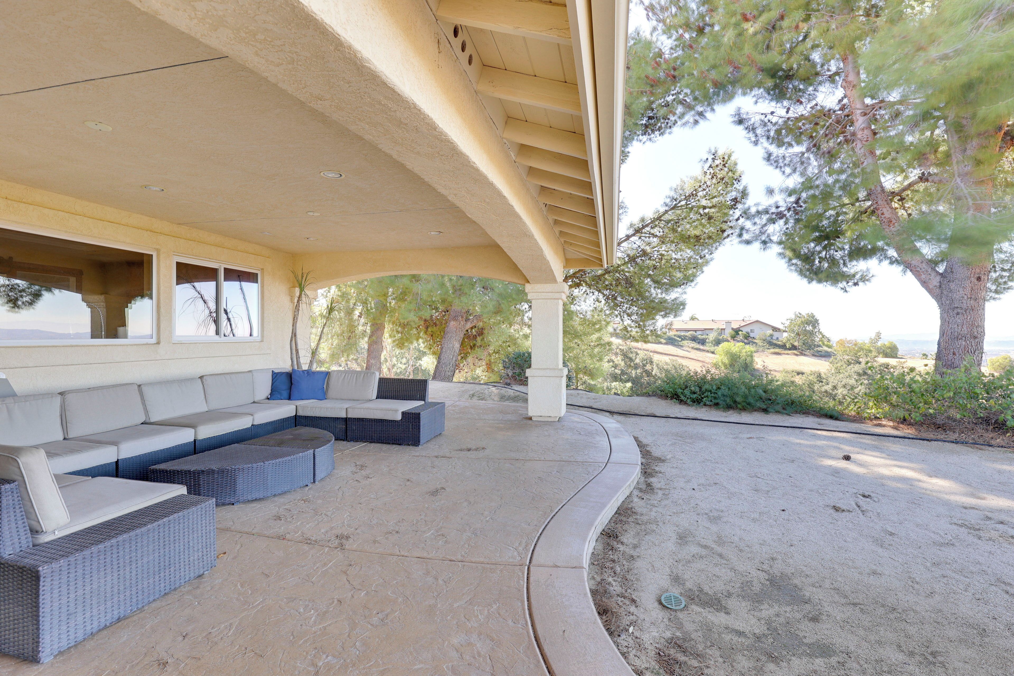 An outdoor covered patio with a long sofa and two side tables, surrounded by greenery and a view of the landscape.
