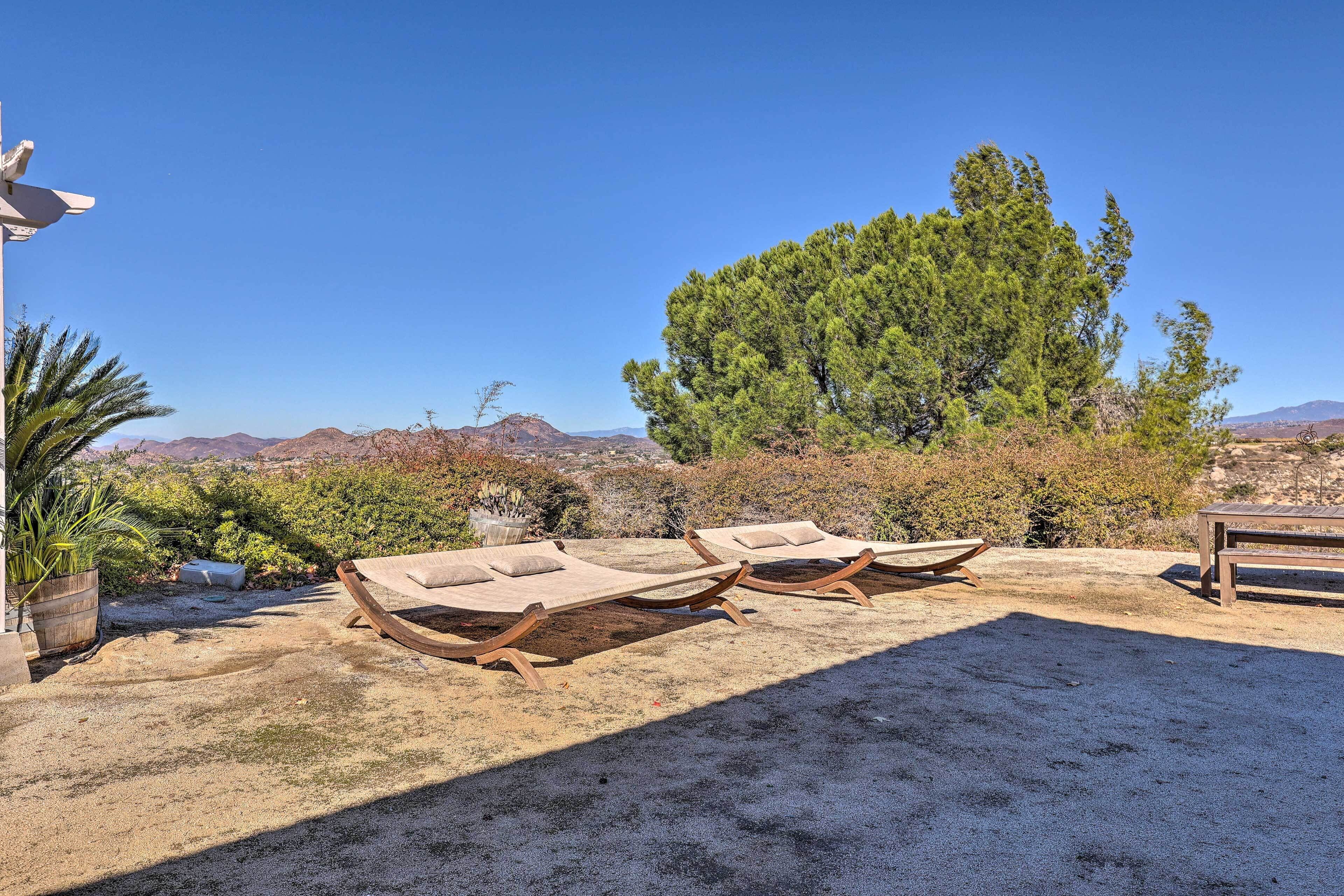 A patio with two wooden lounge chairs and a wooden table, surrounded by a gravel area with a view of mountains and trees.