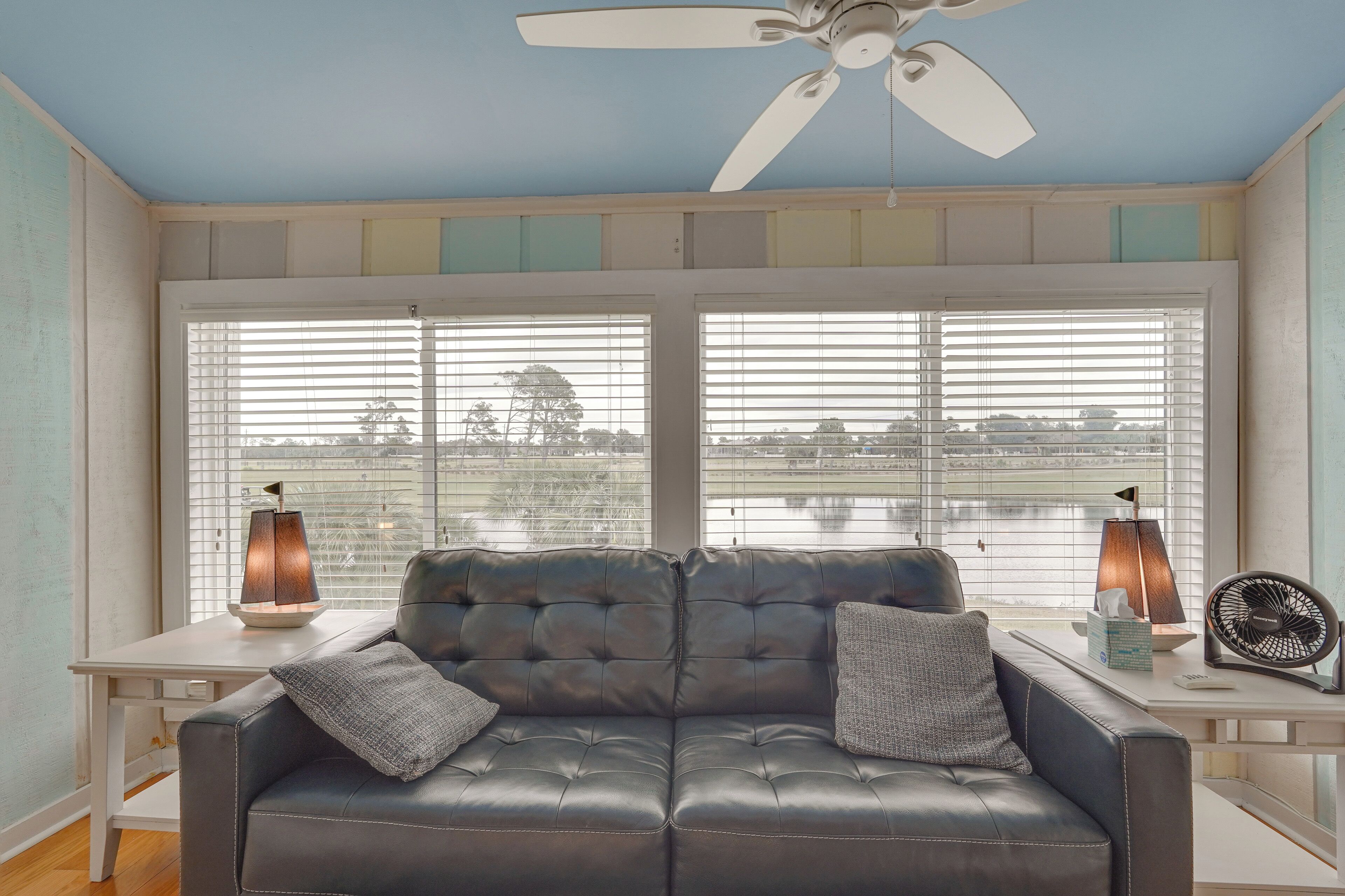 A room with a black leather sofa, two side tables, a ceiling fan, and large windows with blinds.