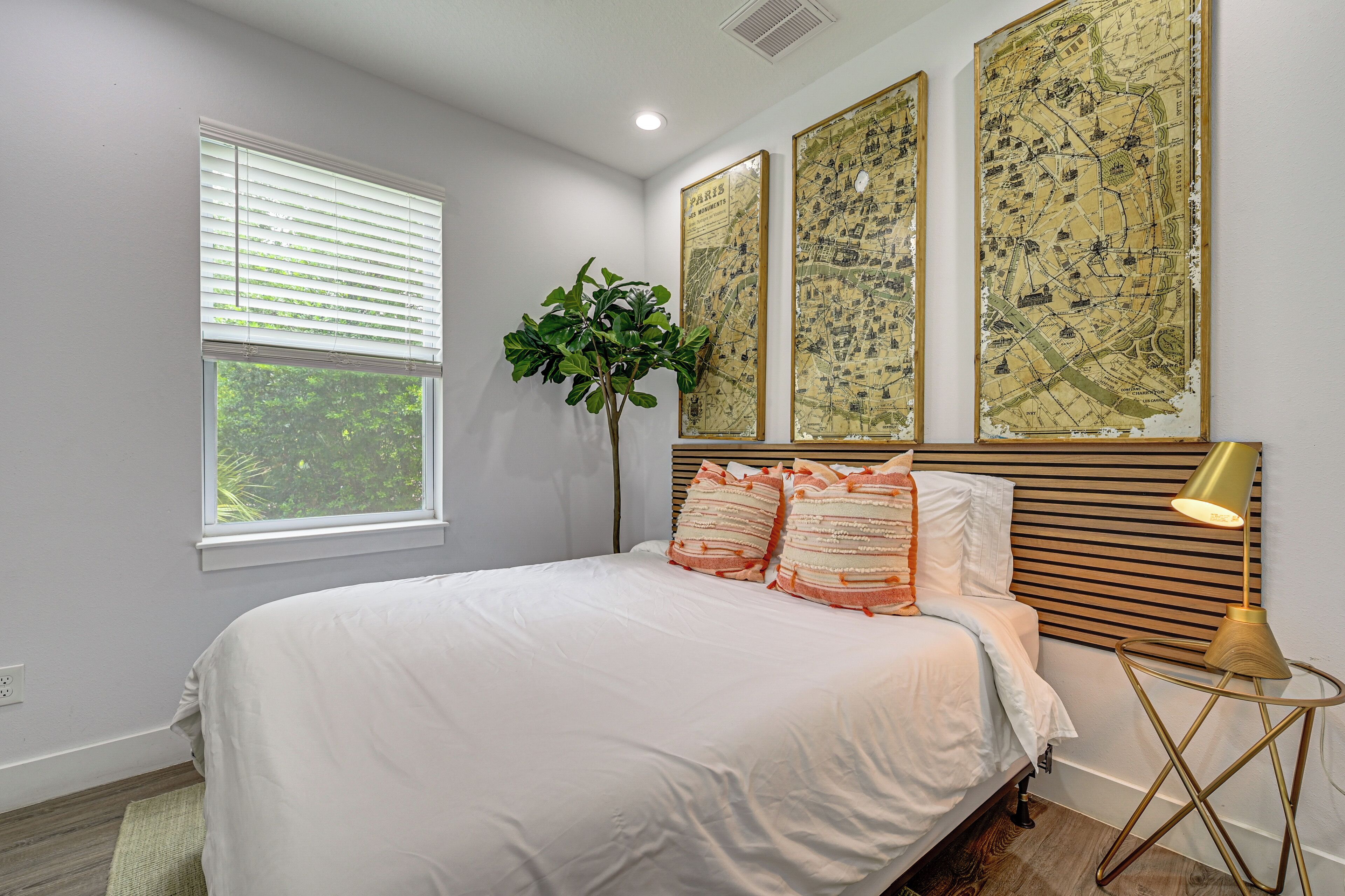 A bedroom with a bed, a window with blinds, a plant, and three framed maps on the wall.