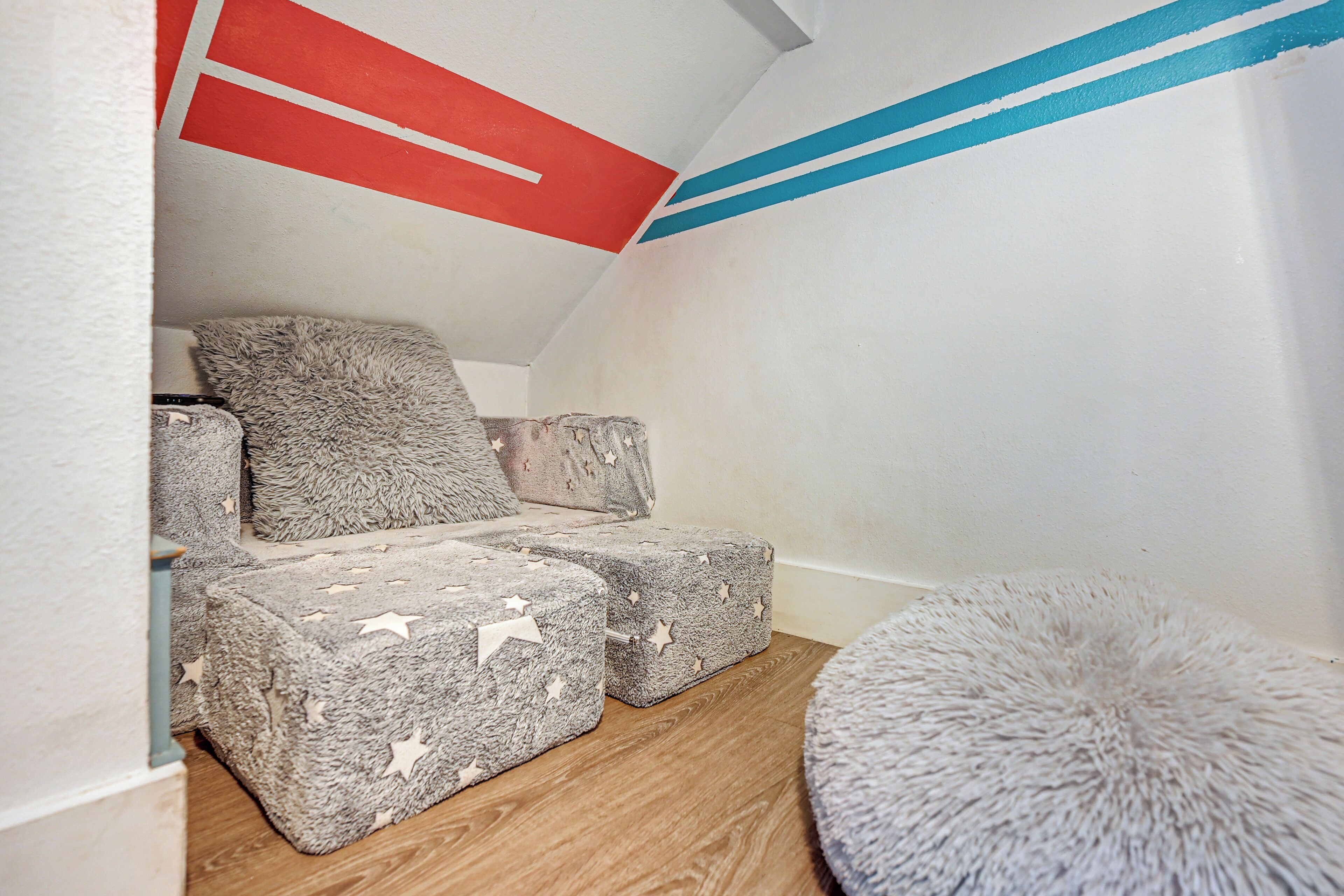 Ein gemütliches Schlafzimmer im Dachgeschoss mit schrägen Wänden, einem flauschigen grauen Teppich und einem kleinen runden Ottomanen.