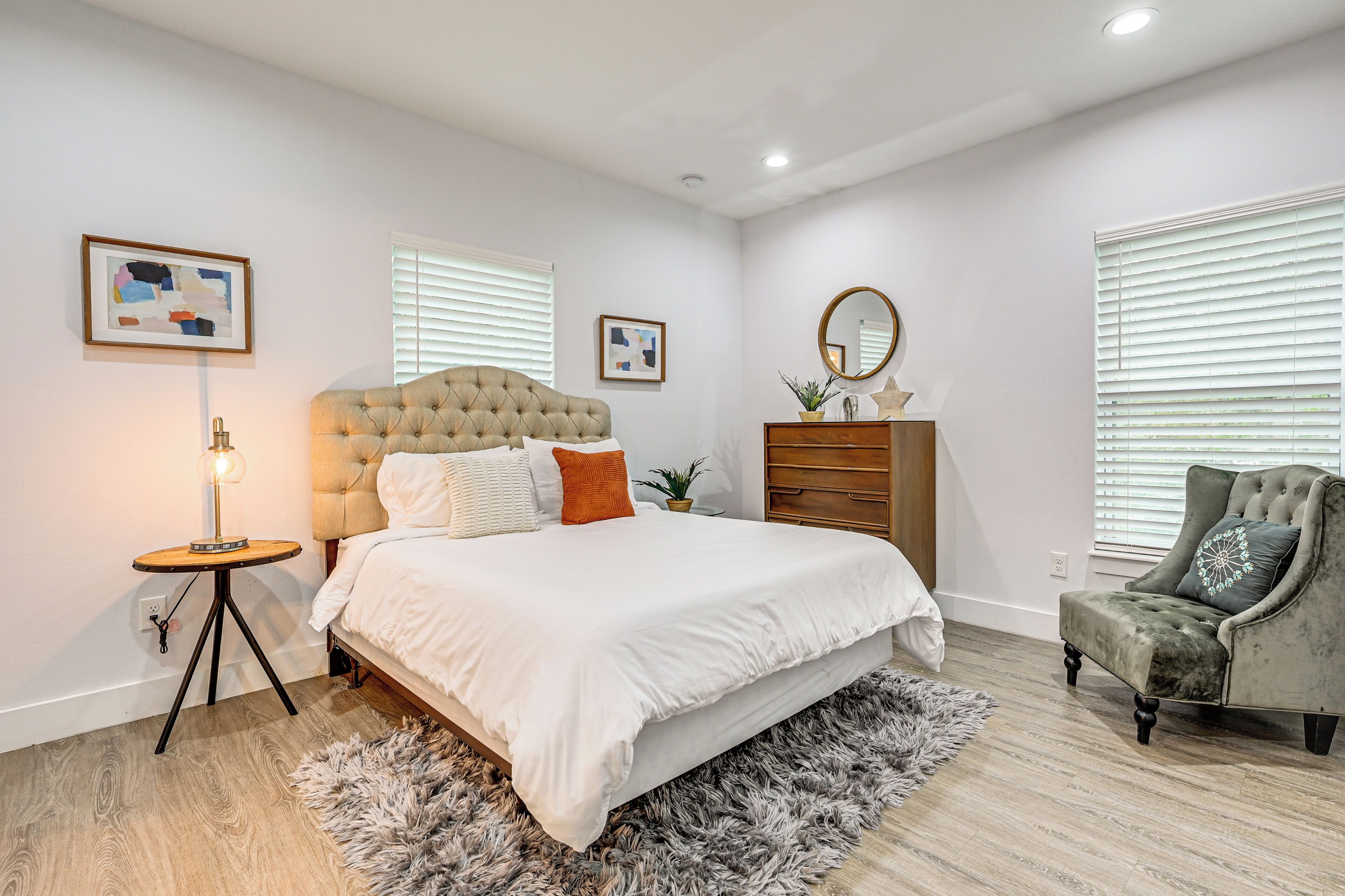Une chambre à coucher avec un lit doté d’une tête de lit capitonnée, une table de chevet ronde, une commode, une chaise et deux fenêtres équipées de stores.