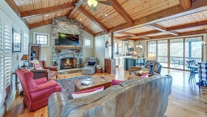 A spacious living room with a stone fireplace, wooden ceiling beams, and a large window overlooking a deck. - Mountain-view Georgia Cabin w/ 2 Decks (Dillard)