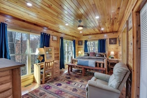 A log cabin interior with a wooden ceiling, large windows with curtains, a bed with a blue blanket, a chair, and a desk with a lamp.