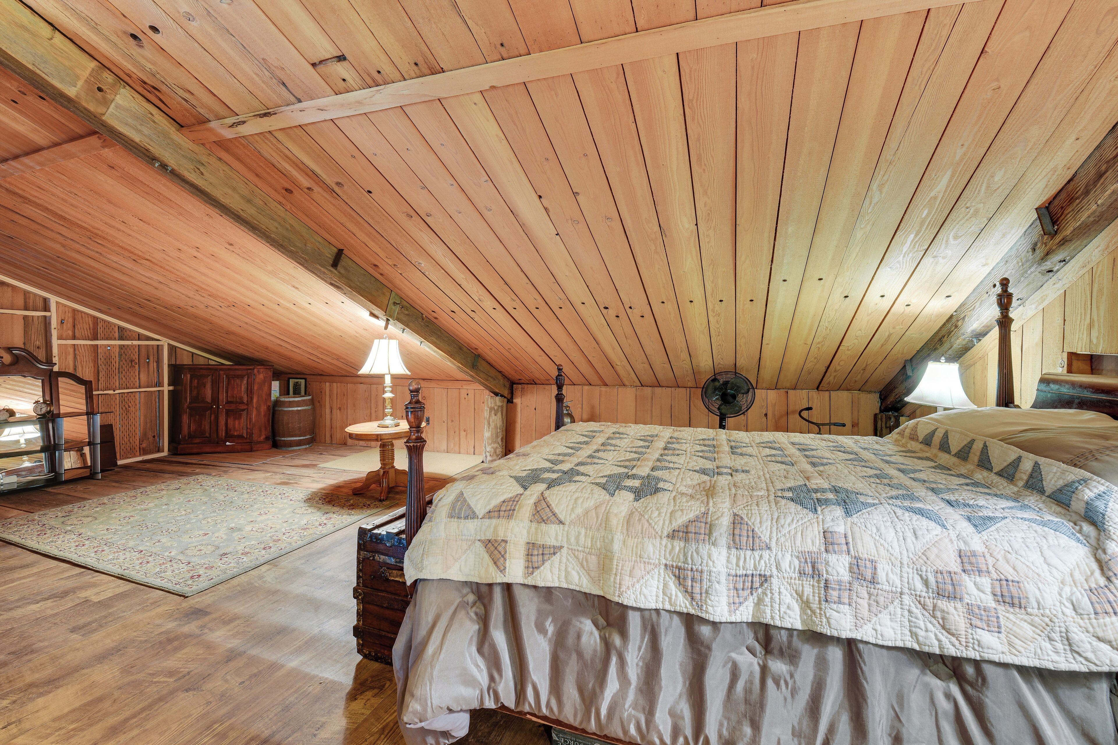 A cozy attic bedroom with a large bed, wooden ceiling beams, a small table, and a fan.