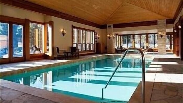 Una piscina interior con agua azul cristalina, vigas de madera en el techo y amplias ventanas a lo largo de las paredes.