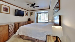 Een slaapkamer met een bed, een plafondventilator, een aan de muur bevestigde televisie en een raam met gordijnen.