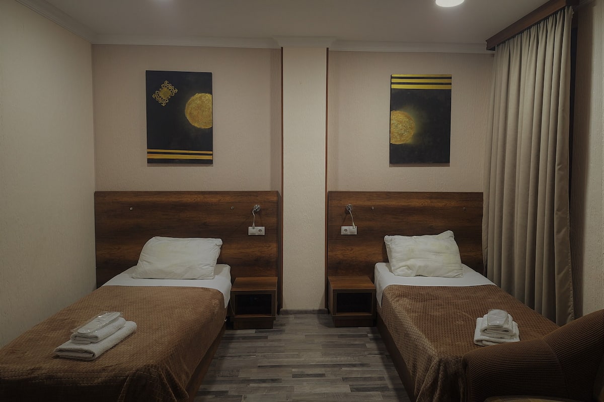 A hotel room with two single beds, wooden headboards, and a small wooden nightstand with a lamp.