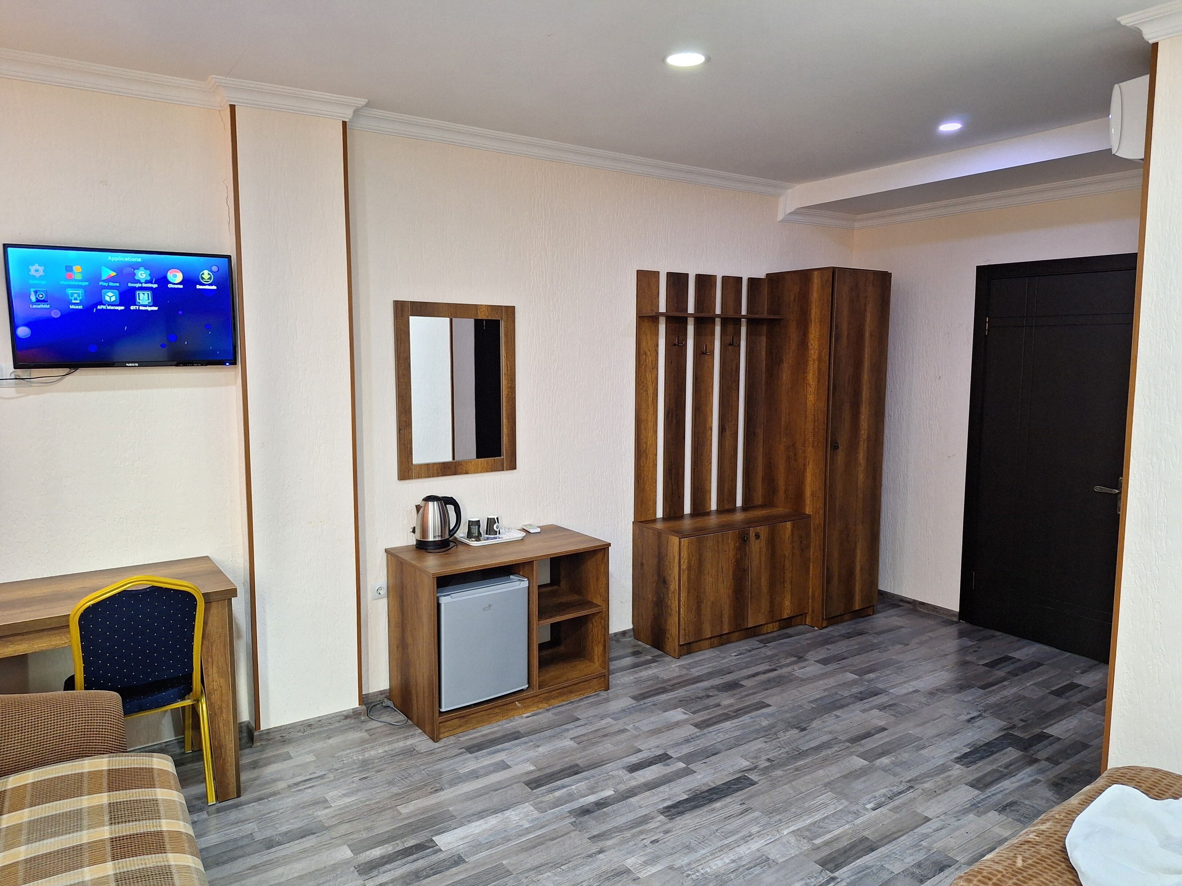 Habitación de hotel con un escritorio de madera, un refrigerador pequeño, un espejo, un armario de madera y un televisor montado en la pared.