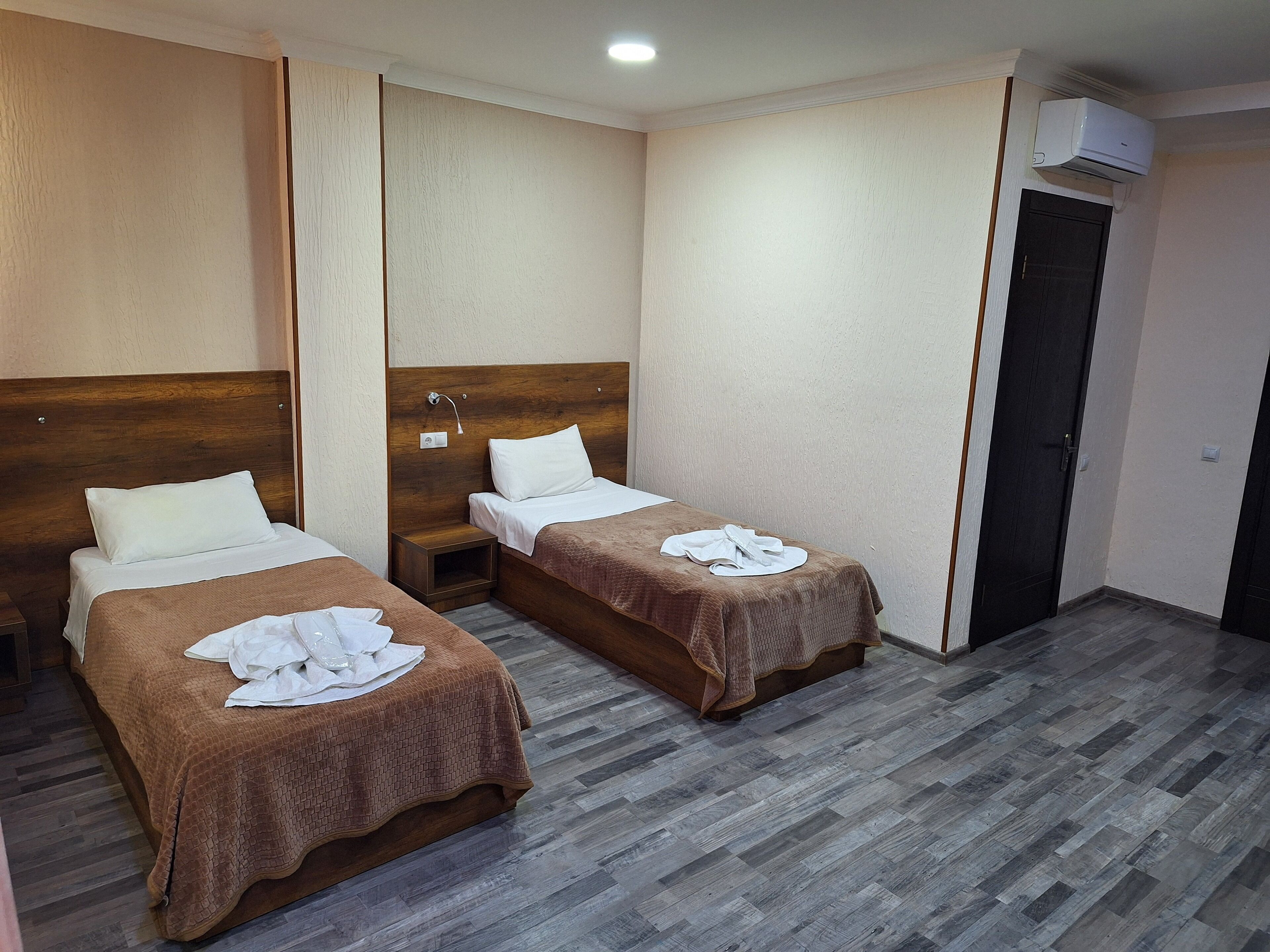 Een hotelkamer met twee bedden, elk met een bruine sprei en witte kussens. Het bed aan de linkerkant heeft een houten hoofdbord, en er is een klein nachtkastje met een lamp. De kamer heeft een houten vloer en een deur aan de rechterkant.