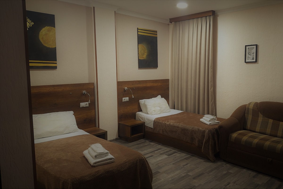A hotel room with two single beds, a sofa, a nightstand, and a wall-mounted lamp.