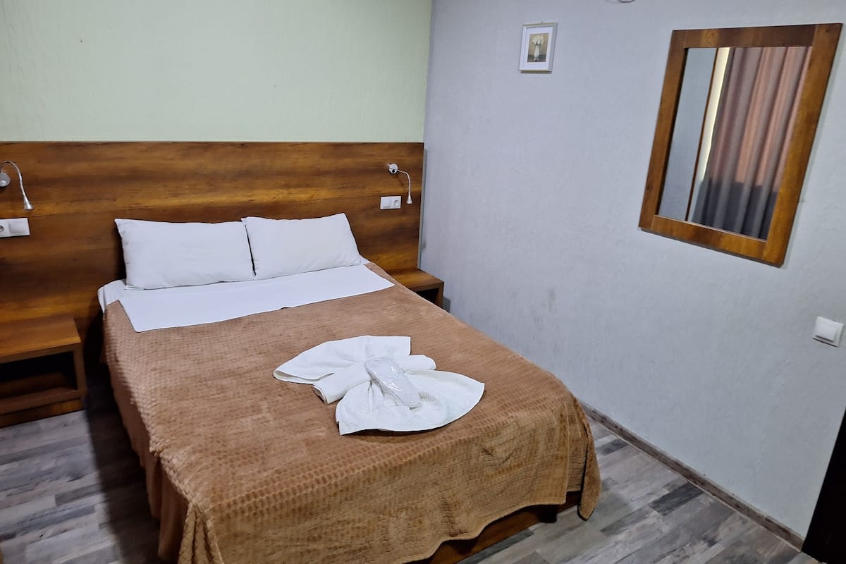 A neatly made bed with a brown blanket and white pillows, a wooden headboard, a bedside table, a wall-mounted mirror, and a small picture frame.