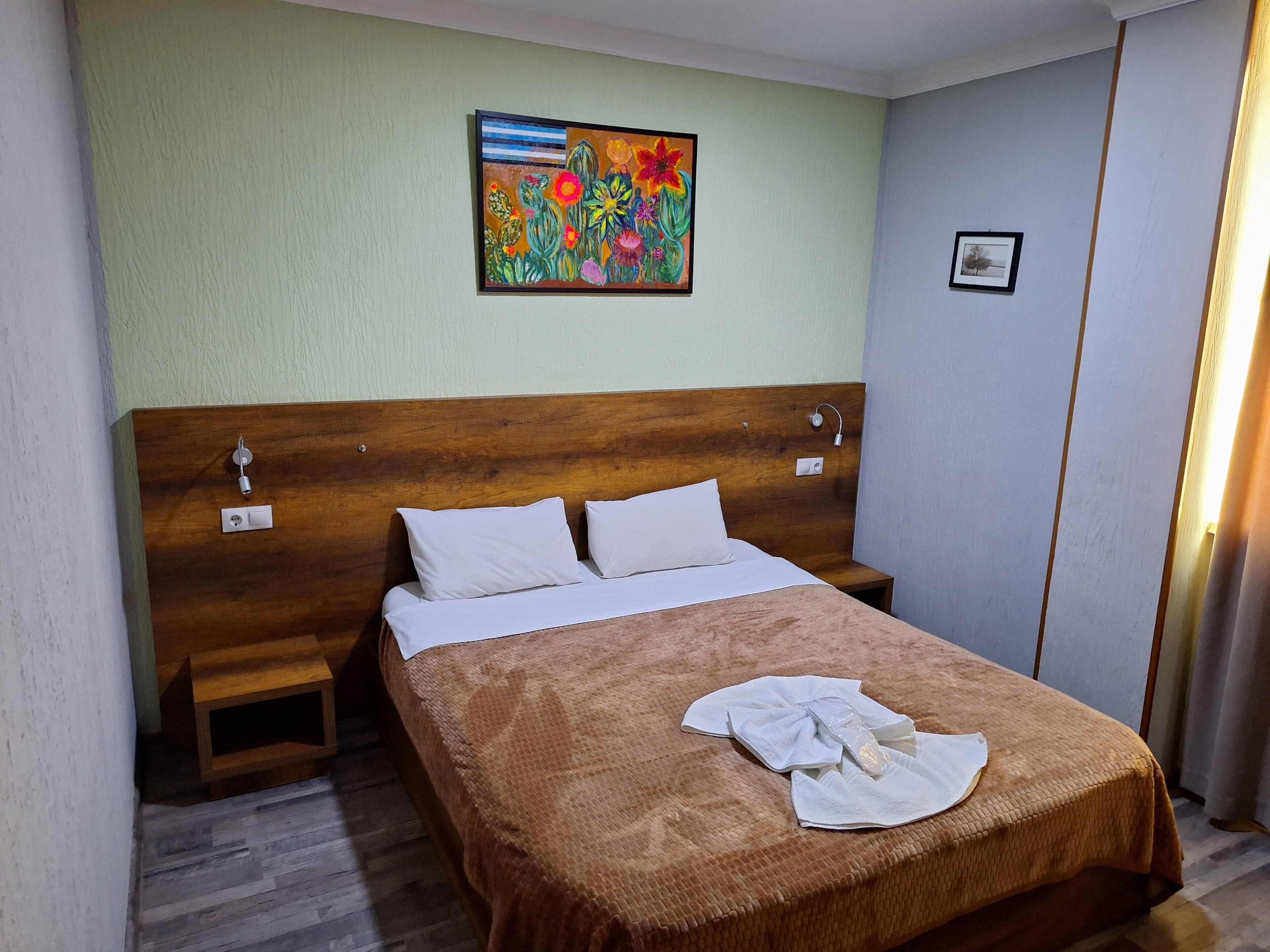 A hotel room with a wooden headboard, a bed with white bedding, a small bedside table, a nightstand, a painting on the wall, and a window with curtains.