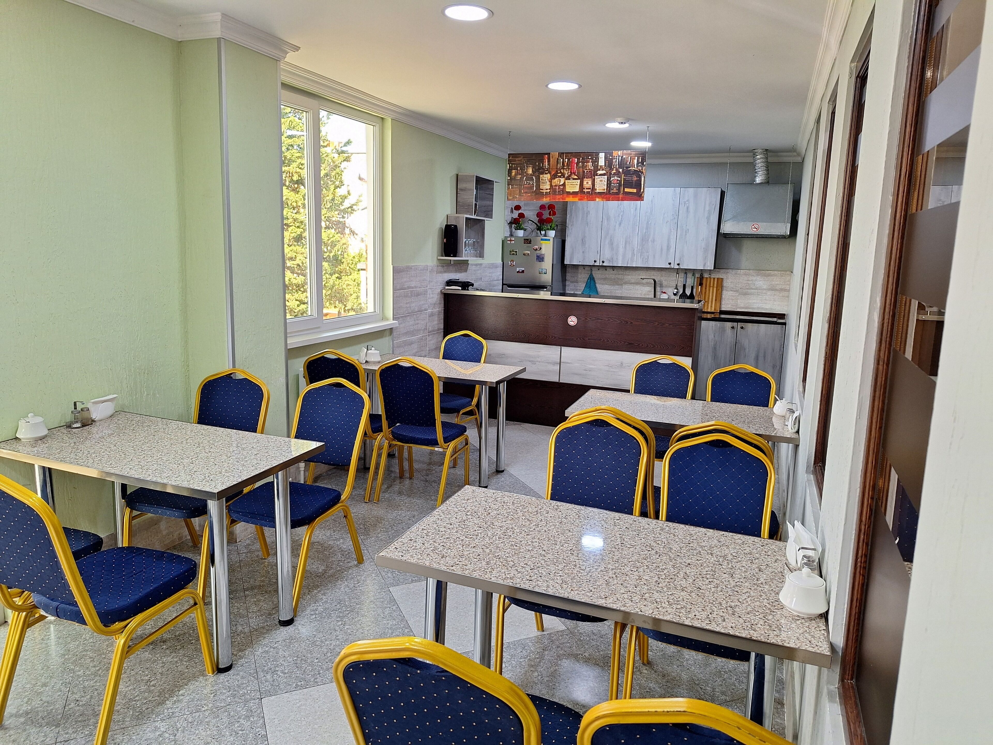 Un espace repas avec des tables et des chaises, une cuisine à l’arrière-plan et une fenêtre donnant sur des arbres.