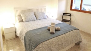 Un dormitorio con cama, una silla, una mesita de noche, un ventanal y dos cuadros en la pared.