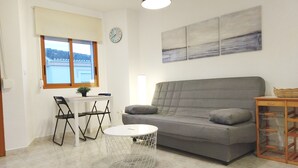 Una sala de estar con un sofá gris, una mesa redonda de centro, una pequeña mesa con sillas, un reloj en la pared y una ventana con persianas.