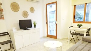 Una sala de estar con un sofá, un televisor sobre un mueble blanco, una mesa de comedor con sillas y un ventanal corredizo que da a un balcón.